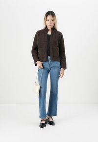 Jeune femme portant une veste zippée marron, un haut noir, un jean bleu, des chaussures noires, tenant un petit sac à main blanc sur fond blanc.