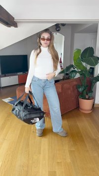 Melnā sporta soma ar "The North Face" logo, valkāta ar gaiši ziliem platām biksēm, baltu garām piedurknēm krekli un pelēkiem slip-on apaviem.