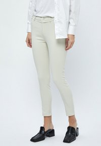 Harmaat, tiukasti istuvat housut, joissa on sileä pinta, keskikorkea vyötärö ja pieni etutasku, yhdistettynä mustiin moolereihin ja valkoiseen nappipaitaan.