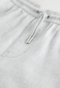 Pantalon de survêtement gris clair avec taille à cordon, fabriqué en tissu texturé et doté d'une seule poche latérale. Embout métallique sur le cordon.