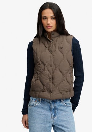 Kvinde iført en brun ærmeløs quiltet vest over en marineblå langærmet sweater og lyseblå jeans, stående foran en hvid baggrund.
