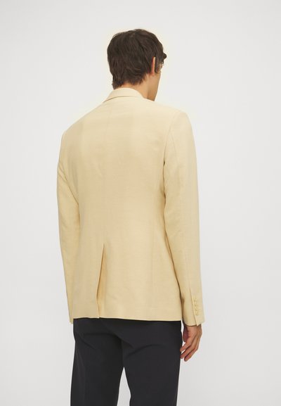 Man in een beige blazer met een centrale rugsplit en zwarte broek, van achteren afgebeeld tegen een effen lichte achtergrond.
