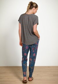 Woman with braided hair wearing a striped short-sleeve top, floral patterned pants, and slippers standing on wooden floor facing away.