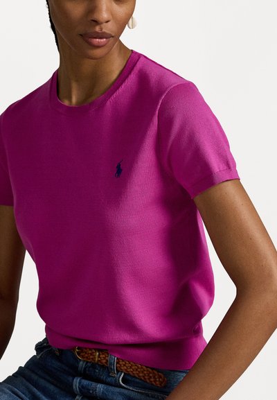 Pull en tricot fuchsia à manches courtes avec un col côtelé et un petit logo bleu sur la poitrine, associé à une ceinture tressée marron et un jean en denim.