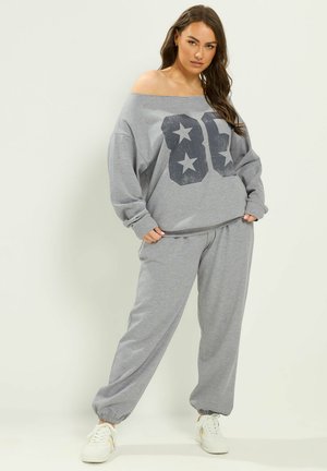 Femme portant un sweat-shirt gris épaules dénudées avec "86" et étoiles, pantalon de jogging assorti et baskets blanches, debout devant un fond uni.