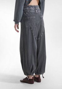 Person trägt locker sitzende, dunkelgraue Hosen mit Gesäßtaschen und Kordelzügen an den Bündchen, kombiniert mit dunkelroten flachen Schuhen, steht auf einem weißen Boden.