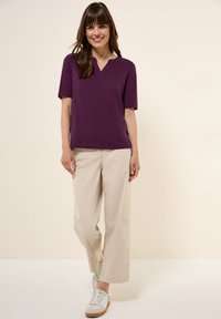 Maglietta viola a maniche corte con scollo a V, abbinata a pantaloni beige a gamba larga e sneakers bianche, su uno sfondo neutro.