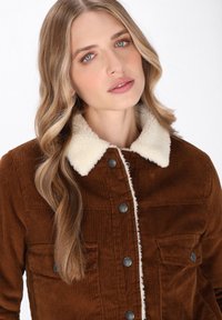 Giacca marrone in corduroy con colletto bianco in pelliccia sintetica, dotata di chiusura a bottoni e due tasche sul petto con bottoni neri.