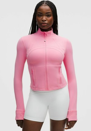 Mujer con trenzas largas que lleva una chaqueta rosa ajustada con cremallera y agujeros para los pulgares, y pantalones cortos deportivos blancos, de pie frente a un fondo liso.