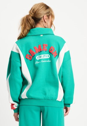 Vrouw met blond haar in een paardenstaart, die een groen-wit sportjack draagt met de tekst „GAME ON ATHLETICS Pom Amsterdam“ op de rug.