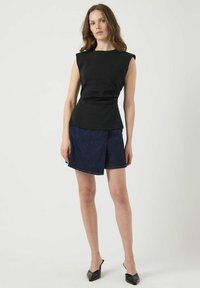 Zwarte mouwloze top met een getailleerde taille, gecombineerd met een donker denim wikkelrok. Het model draagt zwarte puntige muilen.