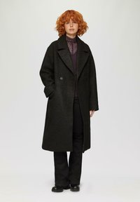 Langer schwarzer Mantel mit strukturiertem Finish und großen Revers, mit einem doppelreihigen Design, getragen über einem dunkel gemusterten Oberteil und schwarzen Hosen.