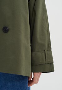 Cappotto verde oliva con ampio colletto, chiusura a bottoni neri e polsini coordinati. Texture liscia con una vestibilità su misura, mostrato insieme a un denim.