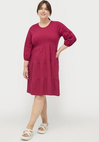 Kurzes, gestuftes Kleid aus tiefroter Baumwolle, mit Rundhalsausschnitt und Puffärmeln, kombiniert mit hellen Kreuzsandaletten.