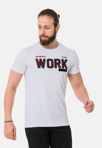Weißes Baumwoll-T-Shirt mit großen schwarzen strukturierten Buchstaben, die "WORK" auf der Vorderseite schreiben, mit zusätzlichem kleinen Text darüber. Kurze Ärmel.