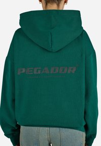 Hoodie oversized de um verde escuro com um logotipo cinza central. Apresenta um capuz espaçoso e punhos canelados, feito de um tecido macio e texturizado.