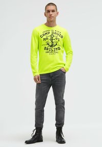 Langärmliges neon-gelbes Hemd mit schwarzem Schriftzug und Anker-Design, kombiniert mit grauer Jeans und schwarzen Schnürstiefeln.