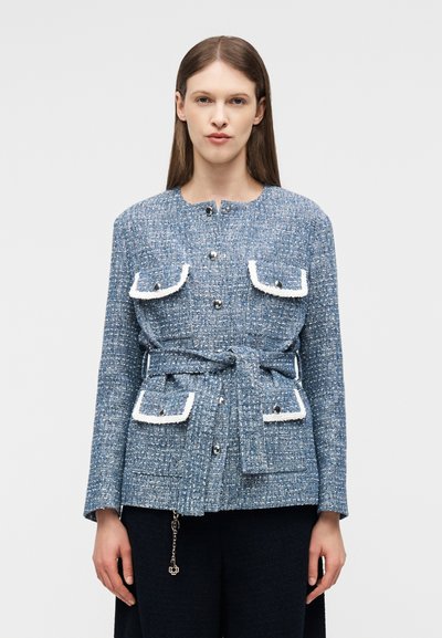 Γυναίκα που φοράει μπλε σακάκι από τουίντ με τσέπες με καπάκι και λευκή επένδυση, ασημένια κουμπιά και ζωνάκι δεμένο στη μέση, στέκεται μπροστά σε απλό φόντο.