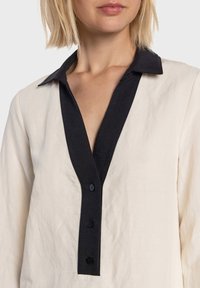 Chemise crème avec un col sombre, dotée d'une patte de boutonnage. Tissu doux et texturé, coupe décontractée avec un décolleté profond en V.