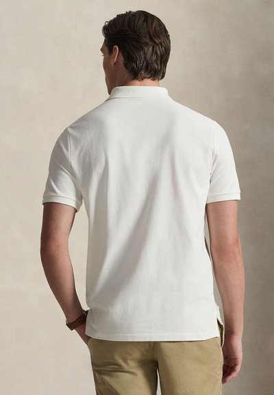 Balta kokvilnas polo krekls ar ribotu apkakli un īsām piedurknēm, ar sānu šķēlumiem un teksturētu apdari. Valkāts kopā ar haki krāsas biksēm.