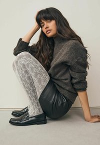 Tmavo hnedý pletený sveter, priesvitné biele čipkové pančuchy s kvetinovým vzorom, čierna kožená sukňa a čierne loafers. Modelka sediaca opretá o stenu.