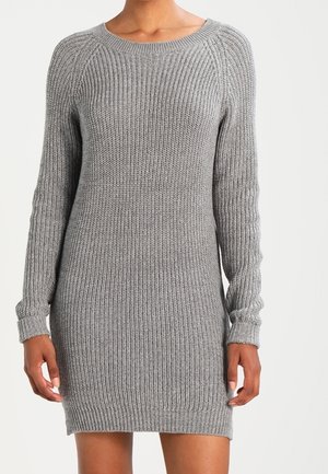 Vrouw draagt een grijze, geribbelde, gebreide trui-jurk met lange mouwen en tot op de knie, tegen een effen lichte achtergrond.