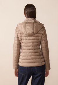 Lys brun polstret jakke med hette, med horisontale quiltede linjer og en glatt, glinsende tekstur. Båret over mørk denim bukser.