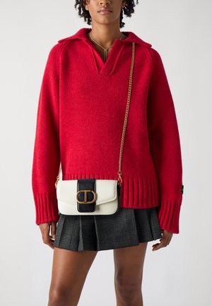 Maglione rosso lavorato a maglia con colletto, caratterizzato da polsini e orlo a costine. Il modello tiene in mano una borsa bianca e nera con accessori dorati.