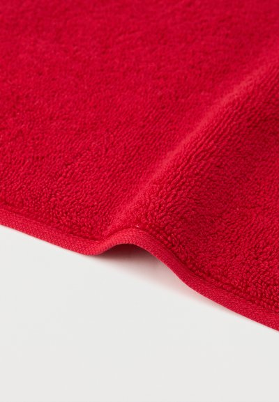 Blødt rødt frottéhåndklædehjørne med en pænt syet kant på en hvid overflade.