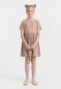 Mlada deklica z blond lasmi, spetimi v dva figi in pletenice, nosi kratko cvetlično obleko s kratkimi rokavi, roza nogavice do kolen in kremaste superge, stoji ob belem ozadju.