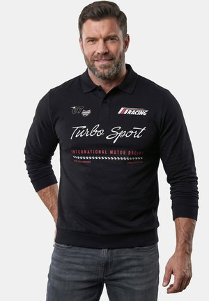 Mand iført en sort langærmet polo med "Turbo Sport" og racertema-logoer, kombineret med blå jeans, stående foran en hvid baggrund.