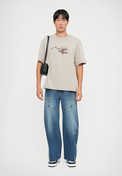 Beige bomull t-skjorte med grafisk trykk og korte ermer, kombinert med vide blå denimbukser og svarte sneakers. Liten svart crossover-veske.