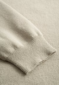 Beige neulottu villapaita hihan ribbineulelahkeella, esitetty vastaavalla tasaisella kangastaustalla.