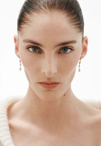 Mujer con el cabello peinado hacia atrás, vistiendo delicados pendientes colgantes y un suave suéter blanco, mirando directamente con una expresión neutral.