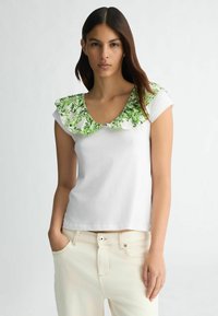 T-shirt blanc à manches courtes avec un col en fleurs vert vif et volanté. Le tissu semble lisse, associé à un pantalon de couleur claire.
