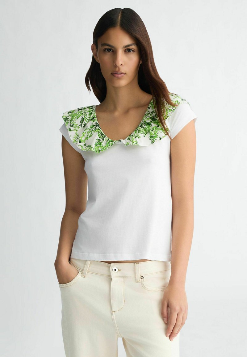 T-shirt blanc à manches courtes avec un col en fleurs vert vif et volanté. Le tissu semble lisse, associé à un pantalon de couleur claire.