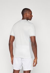 Camiseta atlética blanca de manga corta con espalda de malla texturizada, detalles grises y una línea de color teal en el lateral, combinada con pantalones cortos claros.