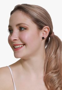 Boucles d'oreilles en stud circulaires noir mat portées par une femme aux longs cheveux ondulés châtain clair, vues de côté sur un fond blanc.