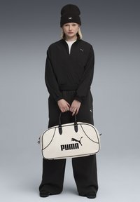 Ung person i sort Puma træningsdragt og hue står og holder en stor hvid Puma sportstaske mod en grå baggrund.