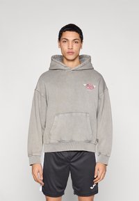 Γκρι φαρδιά φούτερ με κουκούλα, μπροστινή τσέπη και κόκκινο κεντημένο λογότυπο. Συνδυάστε με μαύρα αθλητικά σορτς που έχουν λευκό λογότυπο και λεπτομέρειες.