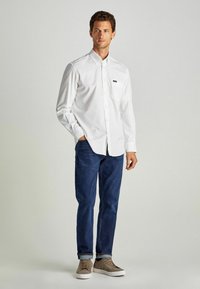 Camicia bianca con bottoni e taschino sul petto, abbinata a jeans blu scuro e sneakers marrone chiaro. Tessuto liscio e vestibilità casual.