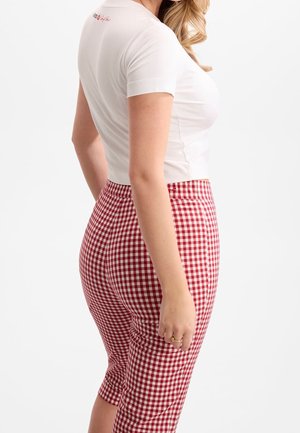Femme portant un haut blanc ajusté à manches courtes et un pantalon taille haute à carreaux rouges et blancs, debout sur un fond blanc.