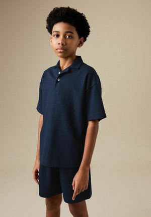 Jongen met kort krullend haar, gekleed in een marineblauw poloshirt en bijpassende korte broek, staand tegen een effen beige achtergrond.