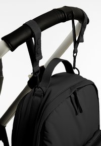 Black backpack hanging from a white stroller handle with black padded grip, secured by two black snap straps and plastic clips.