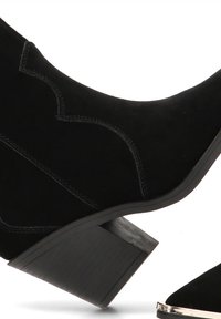 Botas de tobillo de ante negro con un diseño curvado, que presentan un tacón de bloque texturizado y costuras decorativas a lo largo de los lados.