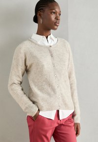 Cardigan en tricot beige avec un col rond, boutons sur le devant et tissu texturé ; associé à une chemise blanche et un pantalon rose.