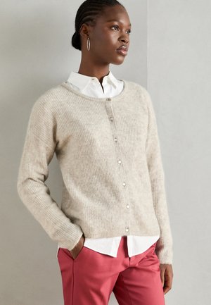 Cardigan en tricot beige avec un col rond, boutons sur le devant et tissu texturé ; associé à une chemise blanche et un pantalon rose.