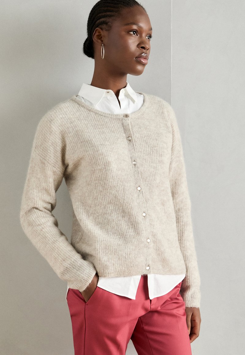 Cardigan en tricot beige avec un col rond, boutons sur le devant et tissu texturé ; associé à une chemise blanche et un pantalon rose.