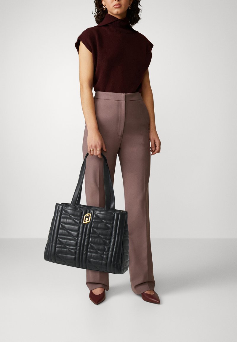 Sac fourre-tout en cuir matelassé noir avec des embellissements dorés, doté d'une poignée supérieure et d'une forme rectangulaire structurée. Associé à un pantalon taupe.