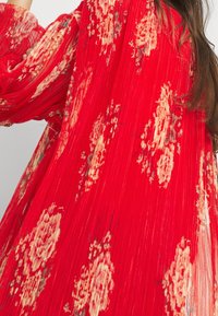 Punainen pleat-kangas toppi, jossa on vaaleanbeige ja kermahohtoinen kukkakuosi. Kankaassa on näkyvä tekstuuri ja muotoillut hihat.